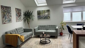 Uma sala de estar moderna com dois sofás, uma mesa de centro e uma mesa de jantar. Há claraboias, uma planta grande e obras de arte nas paredes.