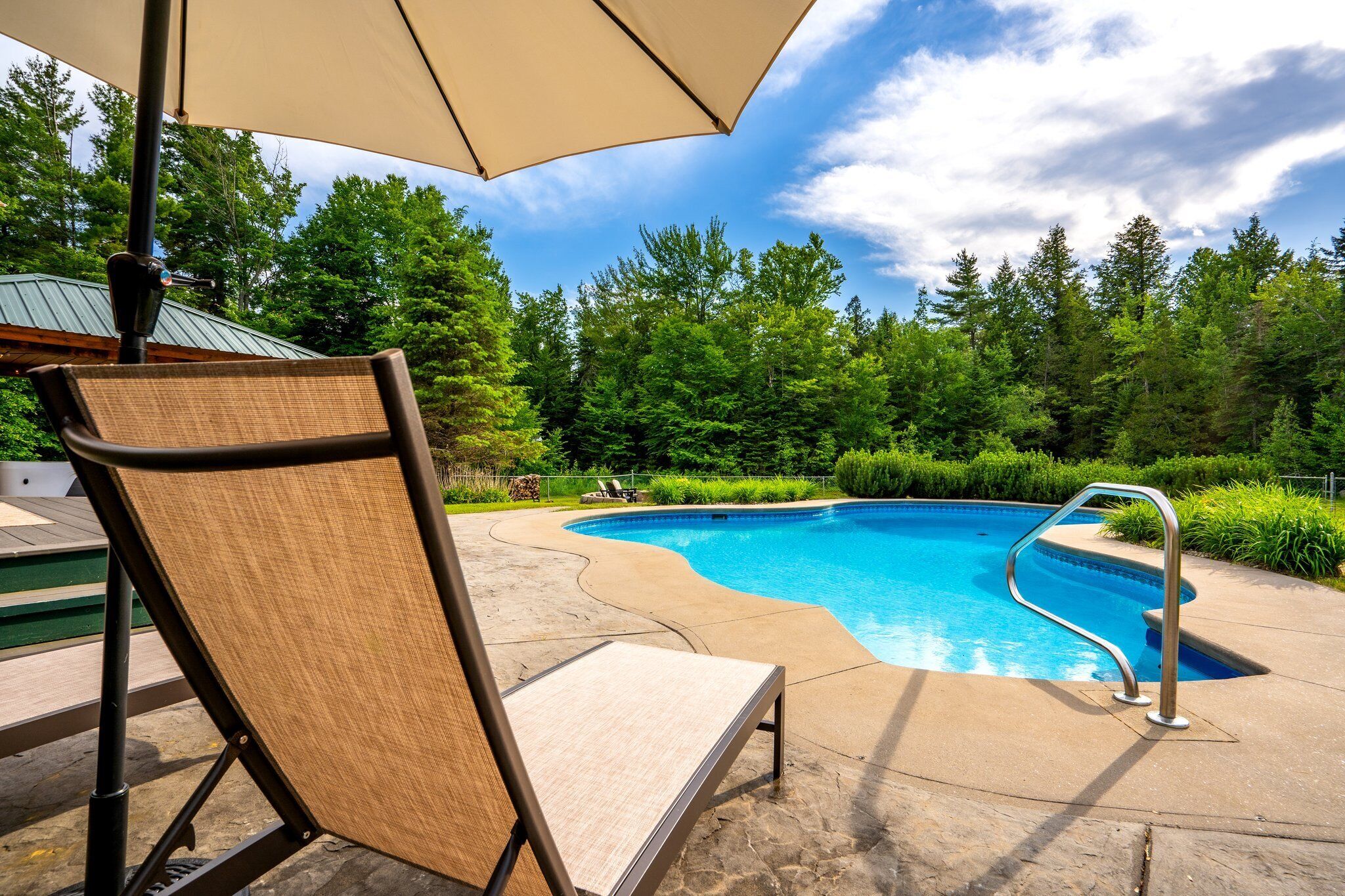 A swimming pool with a metal ladder, surrounded by a concrete deck and a large umbrella providing shade.
