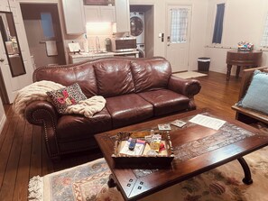 A living room with a brown leather sofa, a wooden coffee table with various items, a kitchen in the background, and a window with curtains. - Cozy vineyard studio with peaceful pond views. (Gilmer)