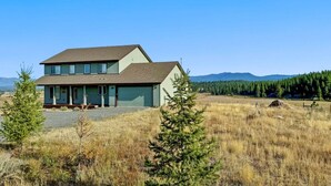 Une maison à deux étages avec un garage, entourée d’un champ herbeux et d’une forêt.