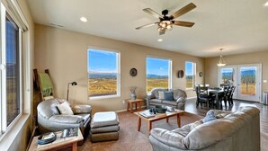 Un salon spacieux avec un grand canapé d’angle gris, un fauteuil en cuir et une table basse. De nombreuses fenêtres offrent une vue sur un paysage rural.