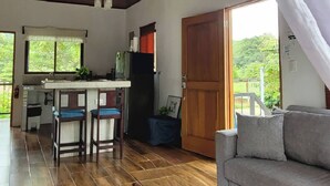 A spacious living area with a wooden floor, a grey sofa, a dining table with two chairs, a kitchen area with a refrigerator, and a large wooden door. - Villa Marina (Nosara)