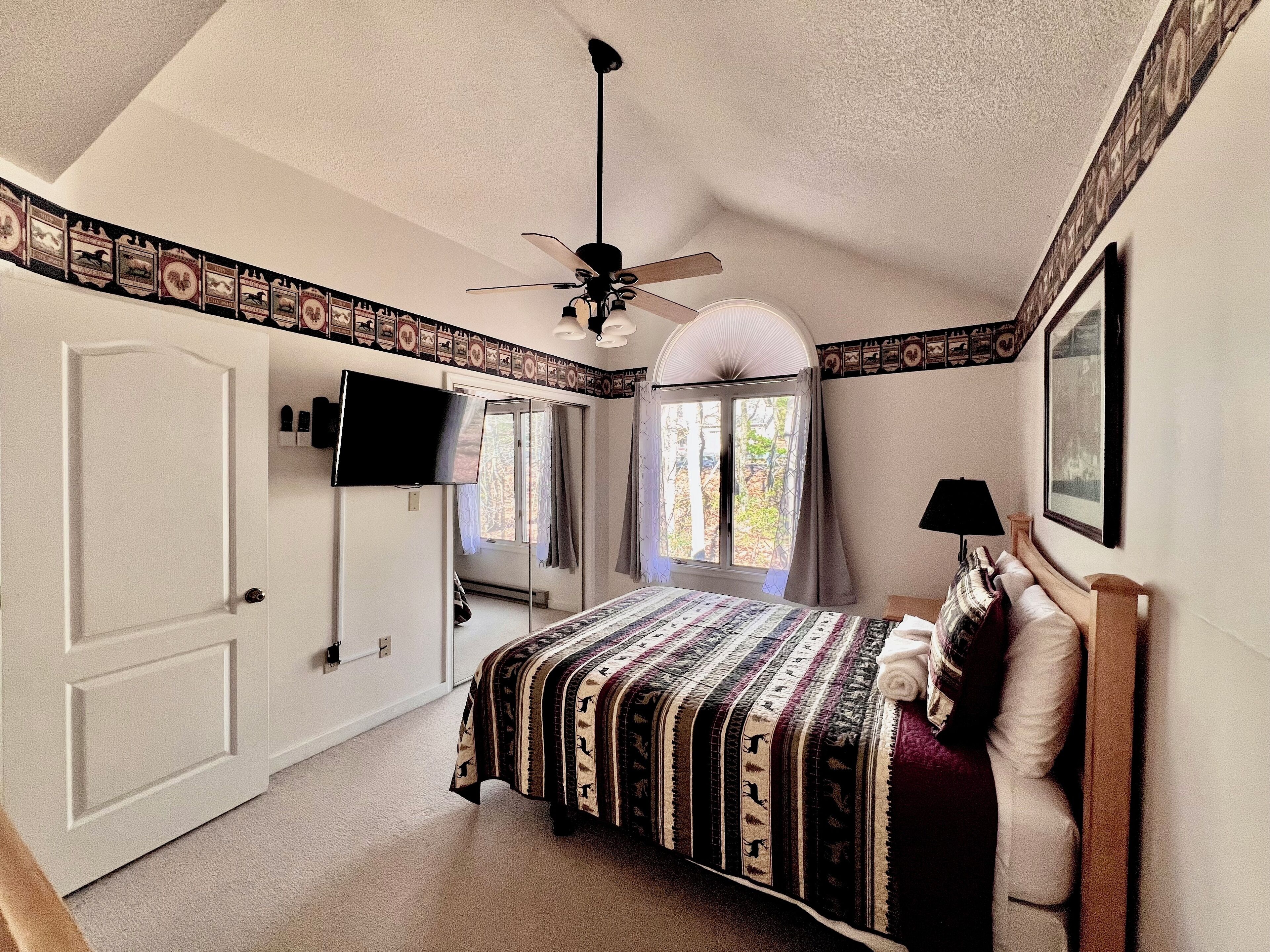 A bedroom with a bed, a ceiling fan, a TV, and a window with curtains.