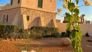 A building with a stone facade and a flat roof, surrounded by a garden with a large decorative stone and flowering plants. - Golden Sand House 2 (Hammamet)