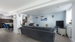 A modern living room with a grey sofa, a dining area with blue chairs, a kitchen with white cabinets, and a TV on a stand. - Offshore -  Carbis Bay (Carbis Bay)