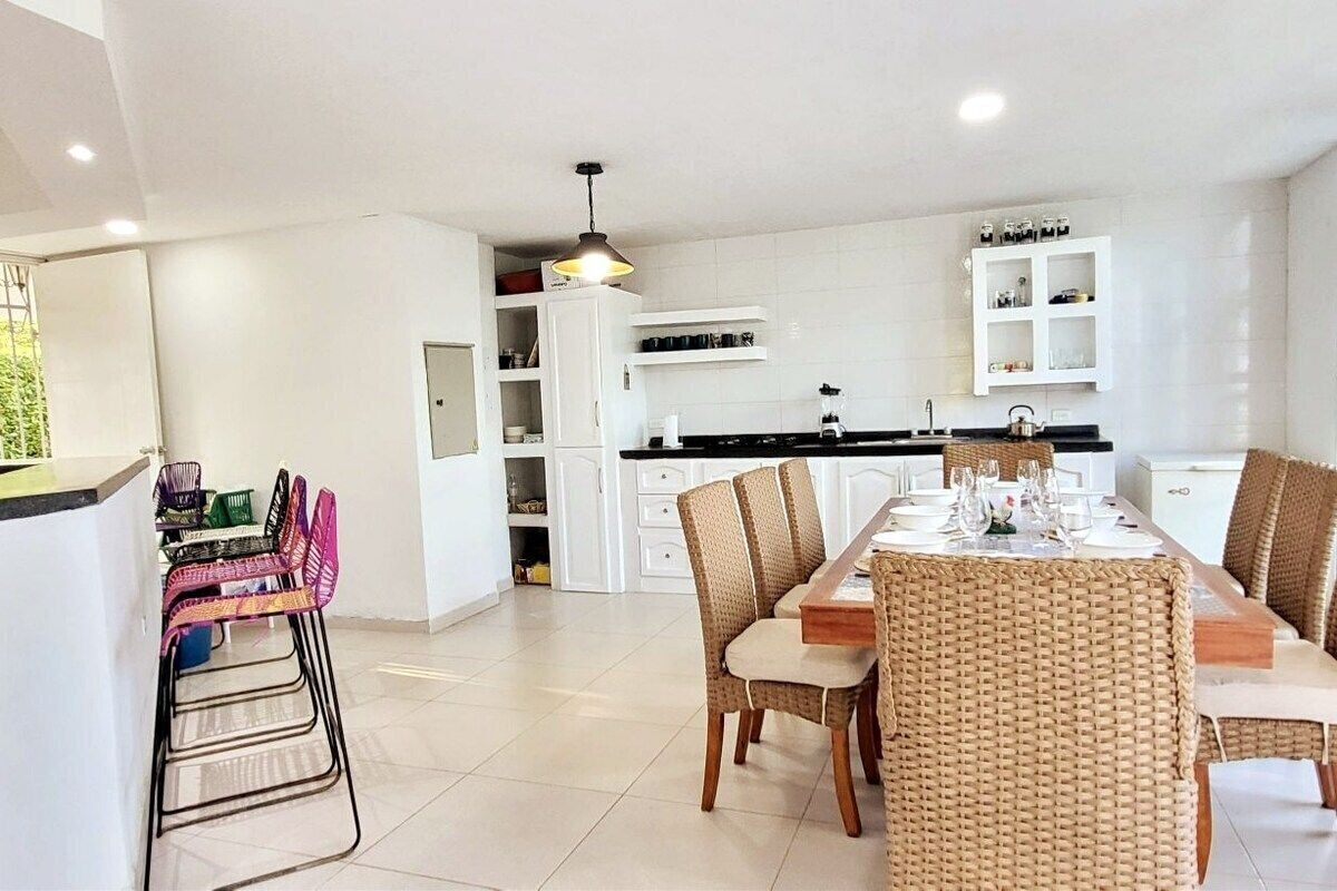 Una cocina moderna con armarios blancos, una zona de comedor con sillas de mimbre y una barra con sillas de colores.