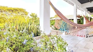 Un hamac sous un pavillon blanc doté d’un sol carrelé, entouré d’une végétation luxuriante et de plantes en fleurs.
