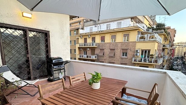 A wooden table and chairs under a large umbrella on a balcony with a city view.