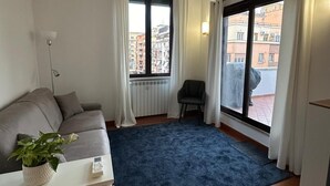 A modern living room with a grey sofa, a small table with a plant, a blue rug, and a chair. There are two windows with curtains, one showing a cityscape view.