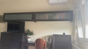 Una habitación con escritorio, silla, armarios oscuros, televisor, planta en maceta y equipo de aire acondicionado.