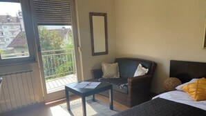 Habitación de hotel con cama, sofá, mesa con papeles, ventana con persianas y vistas a edificios.