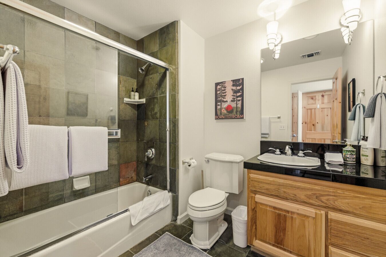 Une salle de bain moderne avec une douche à parois de verre, des toilettes blanches et un meuble-lavabo en bois surmonté d’un plan de travail noir.