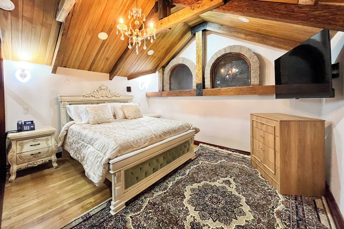 A bedroom with a wooden ceiling, a chandelier, a bed with a patterned bedspread, a nightstand, a dresser, and a large rug.