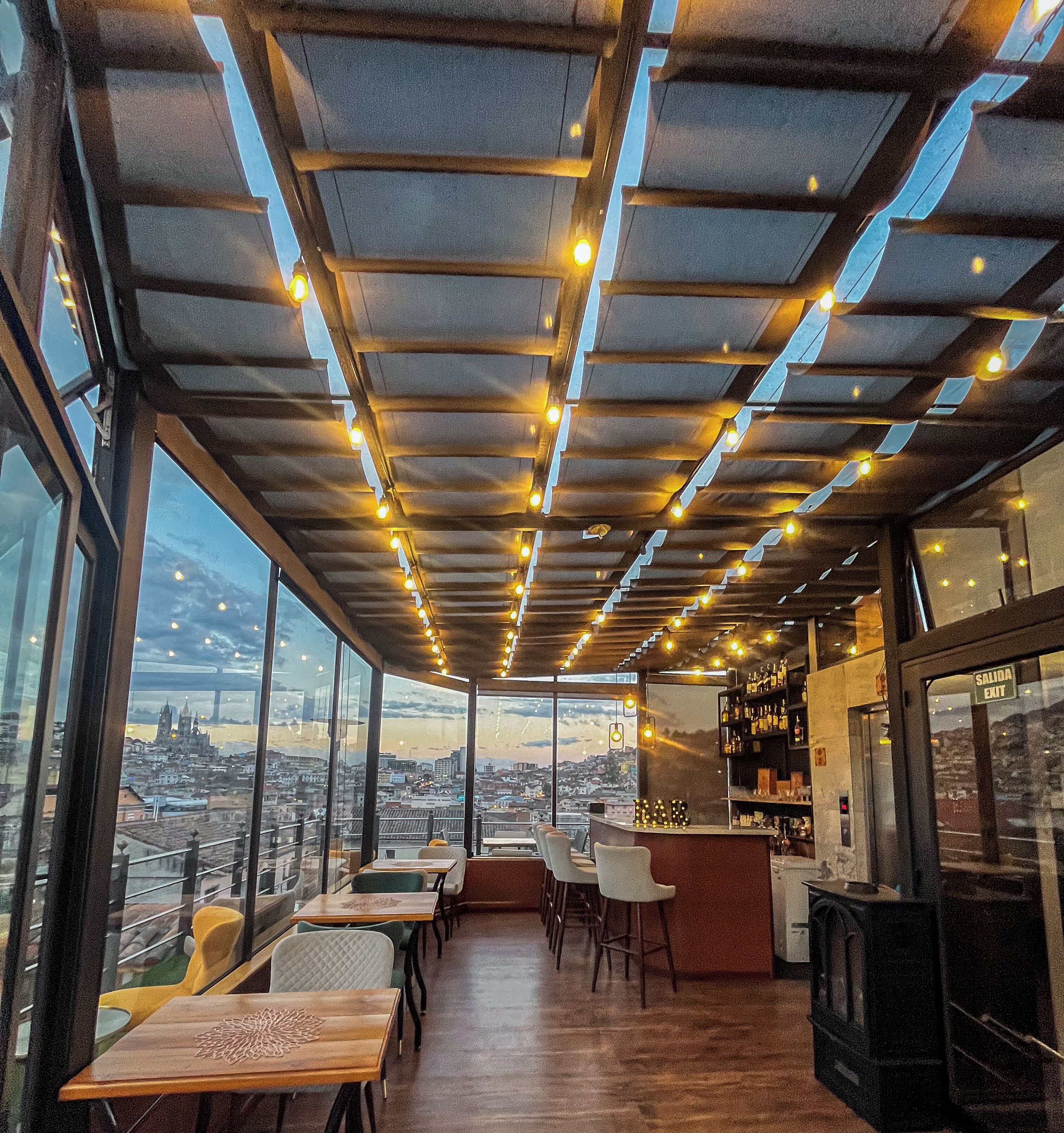 Eine Dachterrasse mit Blick auf die Stadt, Holztische und -stühle sowie eine Bar mit Flaschenregalen.