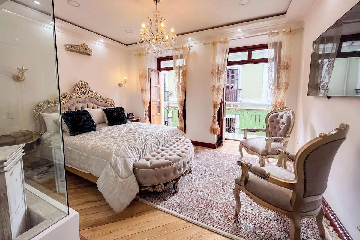 A bedroom with a large bed, a chandelier, a sitting area with a chair, and a view of the outside balcony.