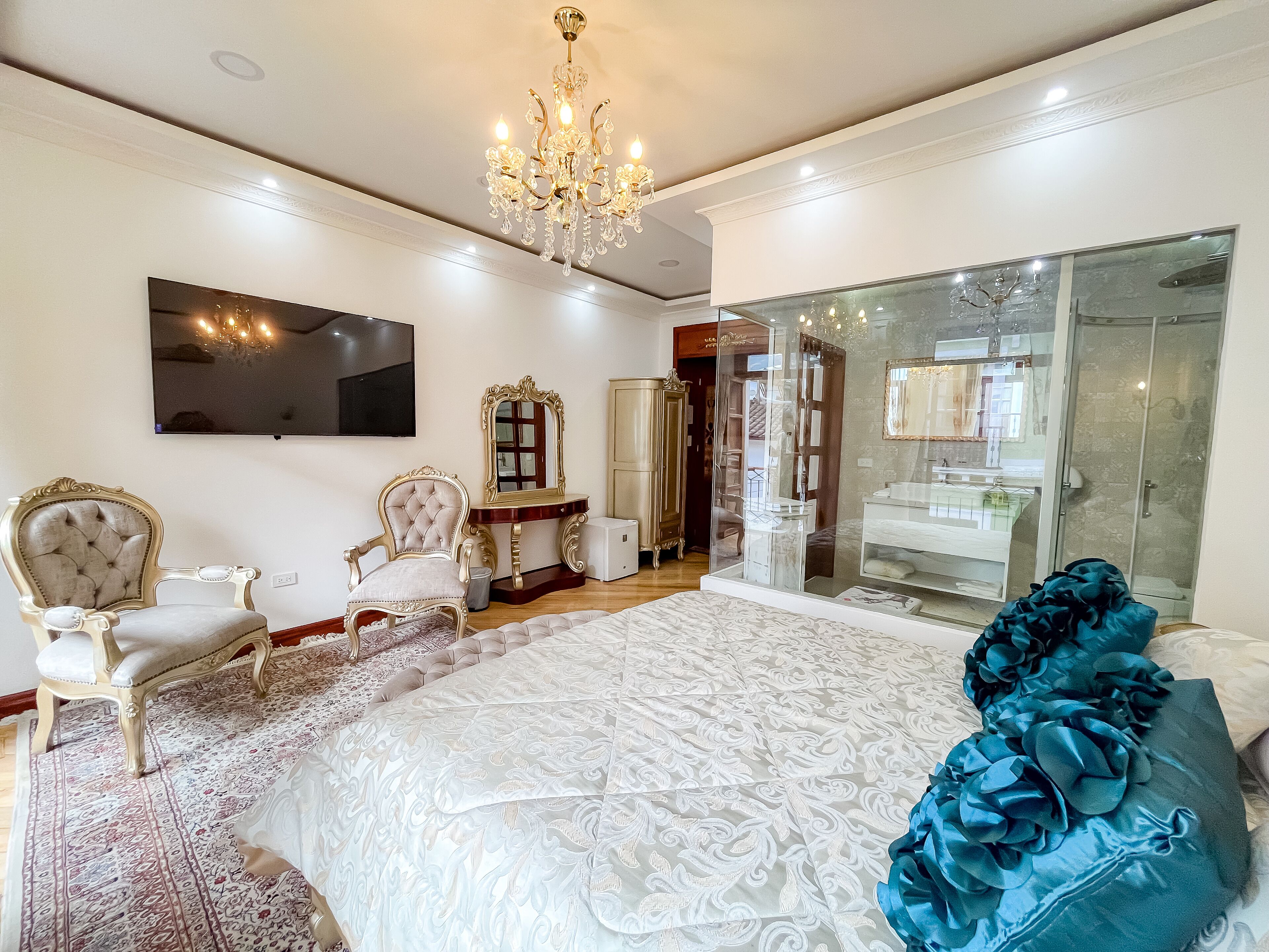 A spacious bedroom with a large bed, two ornate chairs, a chandelier, a TV, and a glass shower enclosure.