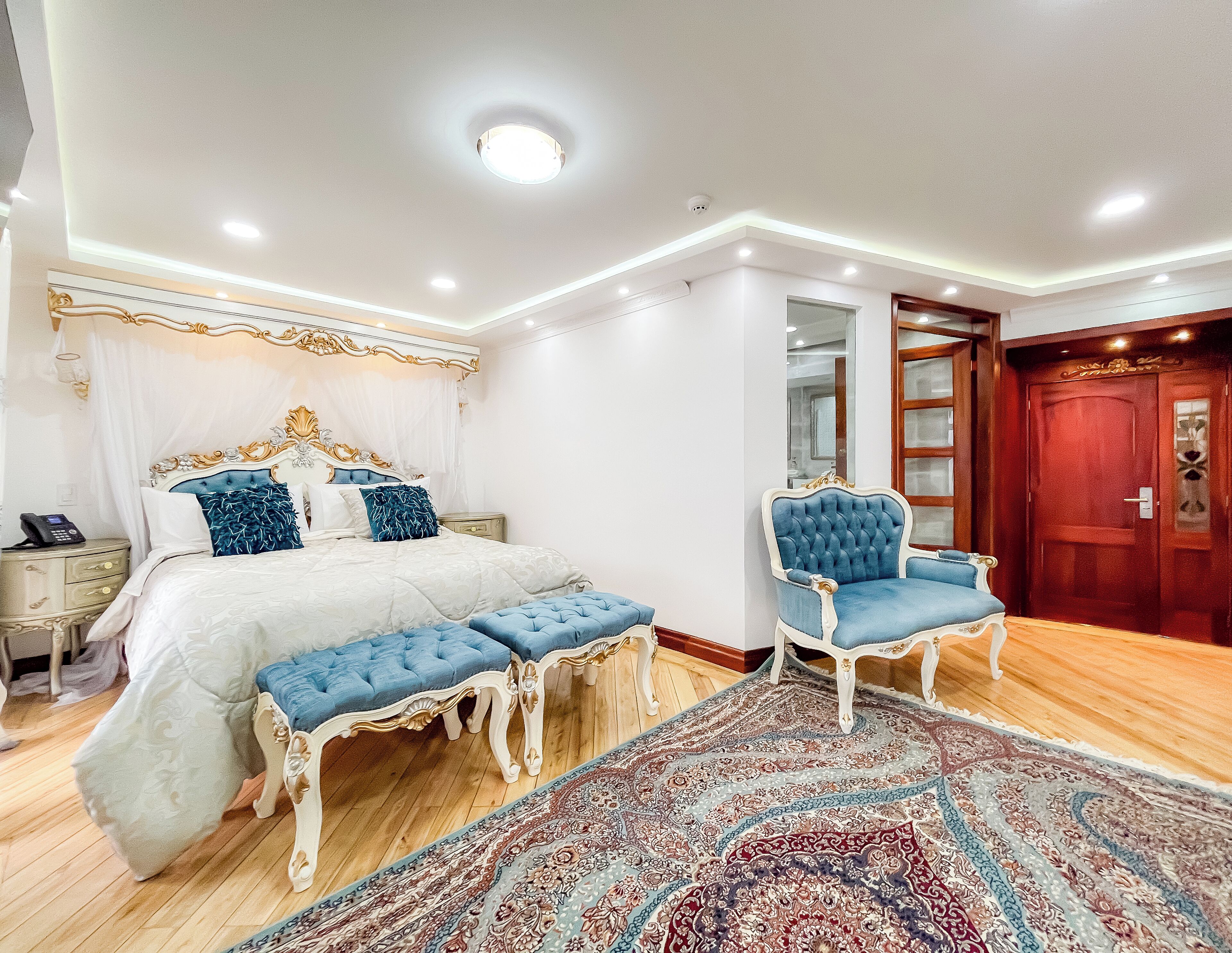 Un dormitorio con una cama grande, una mesita de noche con un teléfono, un sillón azul, suelo de madera y una alfombra estampada.