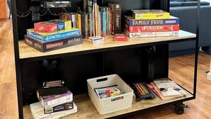 Une bibliothèque contenant divers jeux de société et des livres, une petite table avec du matériel de bureau et un luminaire suspendu.