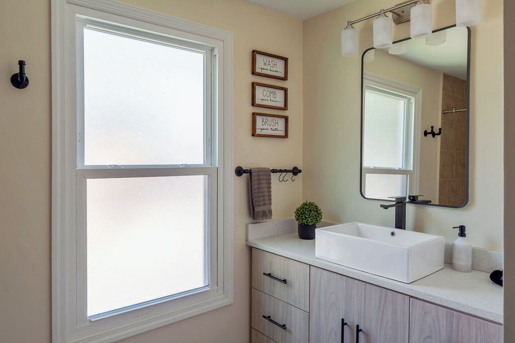 A bathroom with a large window, a sink, a mirror, and three framed signs on the wall.