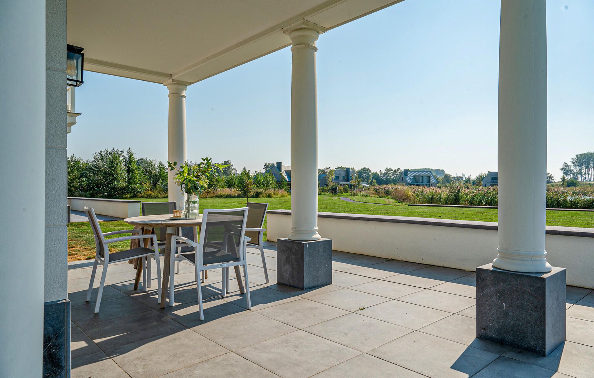 A covered patio with a table and chairs, large columns, and a view of a green landscape and buildings in the distance.