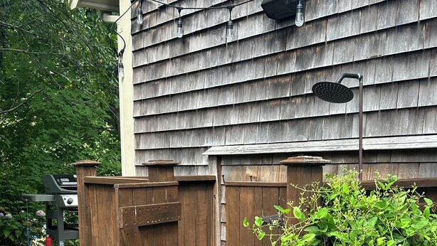 A wooden outdoor shower area with a wooden door, a small wooden deck, and a green plant growing beside it.