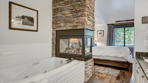 A bathroom with a stone fireplace, a large bathtub, and a bed with a view of trees.
