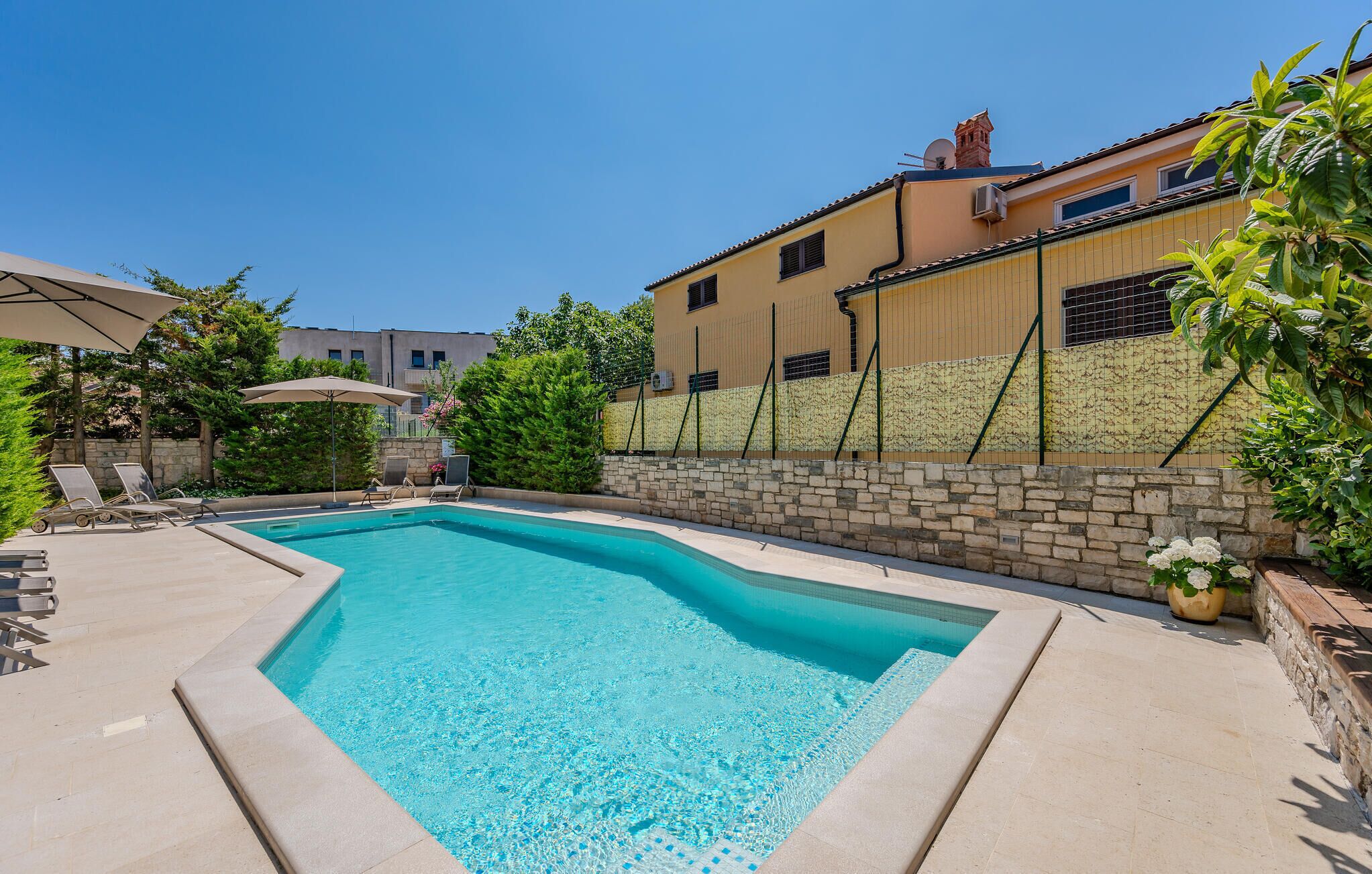 Piscina com espreguiçadeiras e guarda-sóis, cercada por um muro de pedra e vegetação.