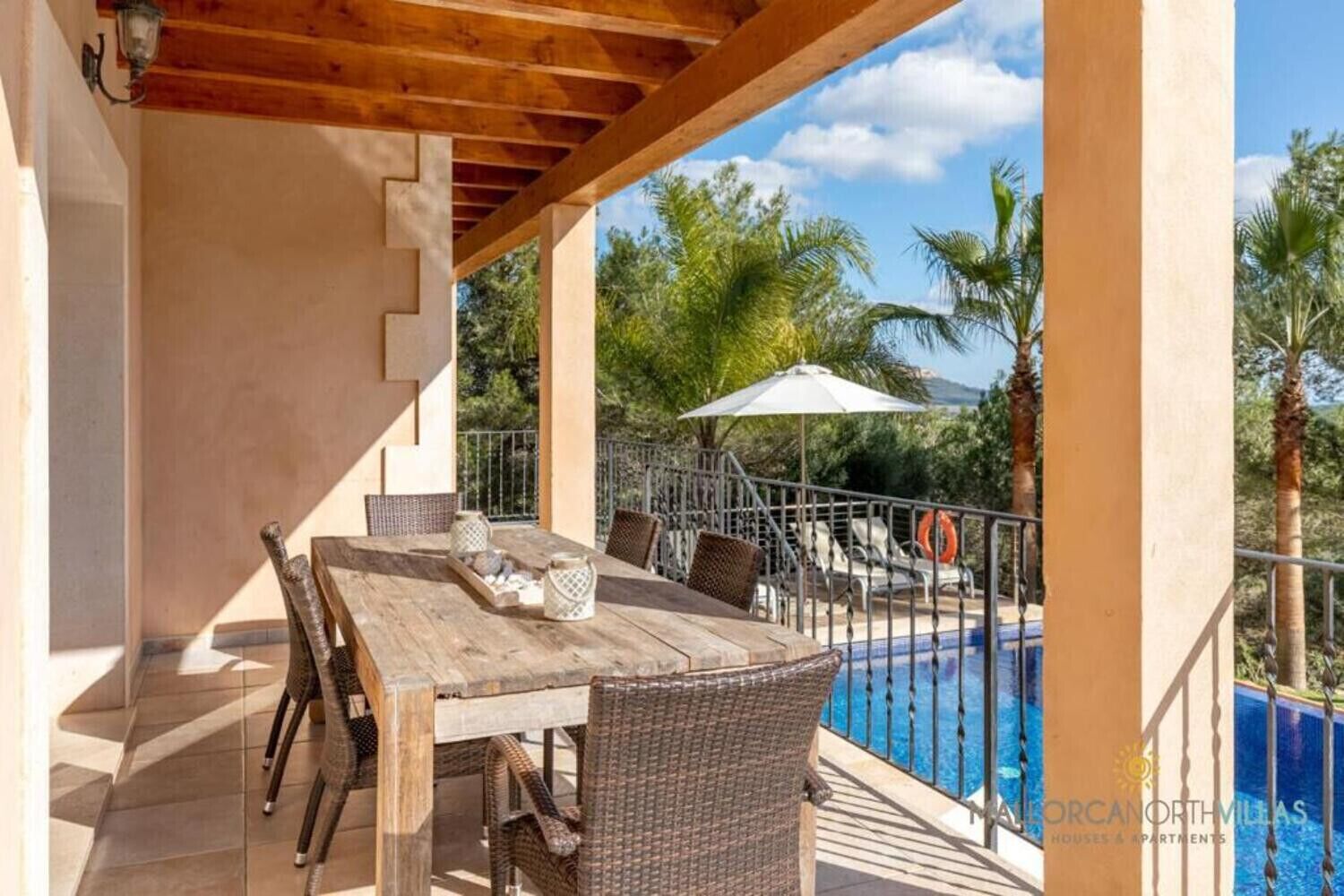 Une terrasse extérieure avec une table en bois et des chaises en osier, donnant sur une piscine bordée de palmiers et surplombant un ciel dégagé.
