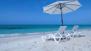 Twee witte lounge stoelen en een witte parasol op een zandstrand met helderblauw water.