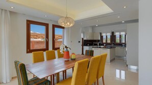Un espace repas moderne avec une table en bois et des chaises jaunes, une cuisine avec des meubles blancs et une vue sur le désert grâce à de grandes fenêtres.