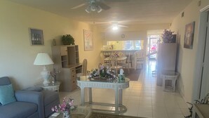 Un salon comprenant un canapé, une table basse, une table avec un plateau en verre orné d’objets décoratifs, un ventilateur de plafond et une vue sur la cuisine.