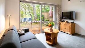 A modern living room with a sofa, a wooden cabinet, a TV, and a sliding glass door leading to an outdoor area with a table and chairs. - Hameau du courant by Interhome (Mimizan)