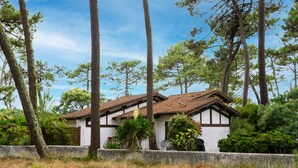 A house with a tiled roof, surrounded by tall trees and a stone wall. - Hameau du courant by Interhome (Mimizan)