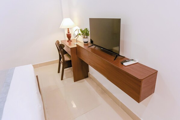 A modern hotel room with a wooden desk, a flat-screen TV, a lamp, a mirror, and a potted plant.
