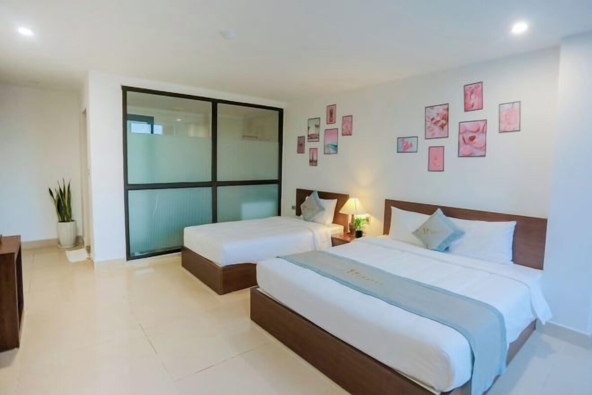 A hotel room with two beds, a sliding glass door, a bedside table with a lamp, and several framed pictures on the wall.