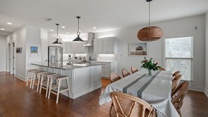 A modern kitchen with white cabinetry, a central island, and bar stools. There is a dining area with a table and chairs. - Waterfront Fairhope Home / Private Pier, Bayfront Views, Screened in Porch (Fairhope)