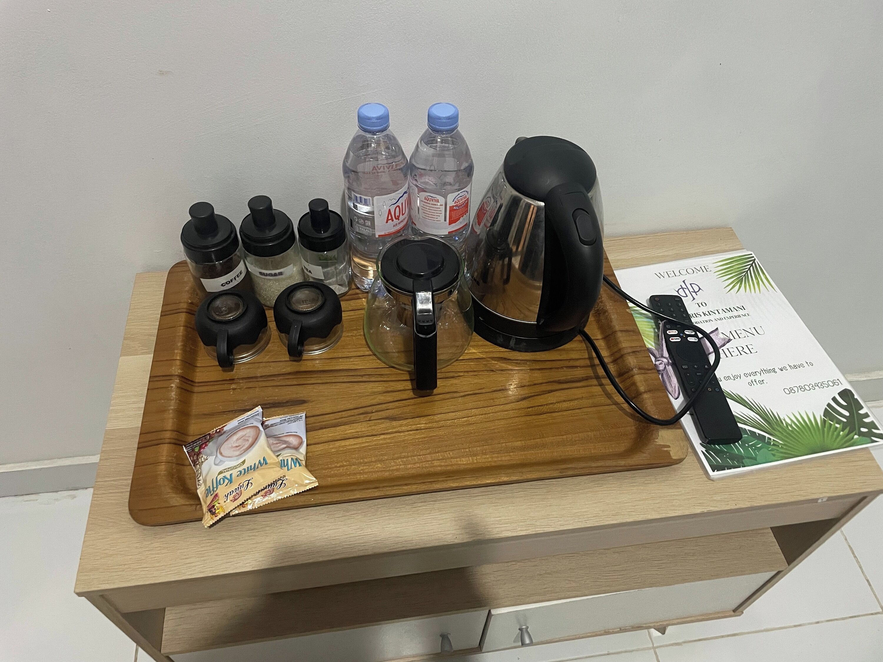 A wooden tray with coffee beans, sugar, water bottles, a kettle, a packet of snacks, and a remote control on a table.