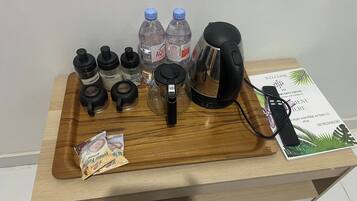 A wooden tray with coffee beans, sugar, water bottles, a kettle, a packet of snacks, and a remote control on a table.