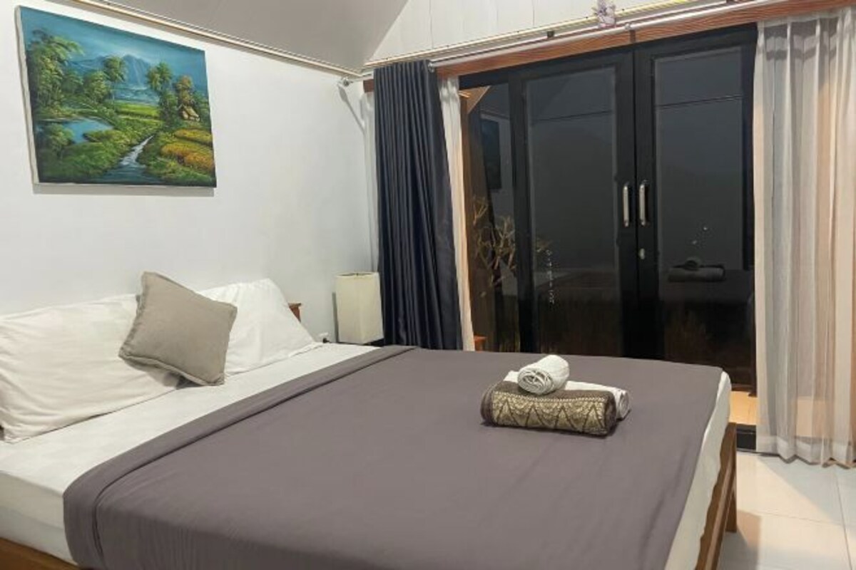 A neatly made bed with a brown bedspread, a white pillow, and a rolled towel. There is a painting of a landscape on the wall, and a dark-colored wardrobe with a glass door.