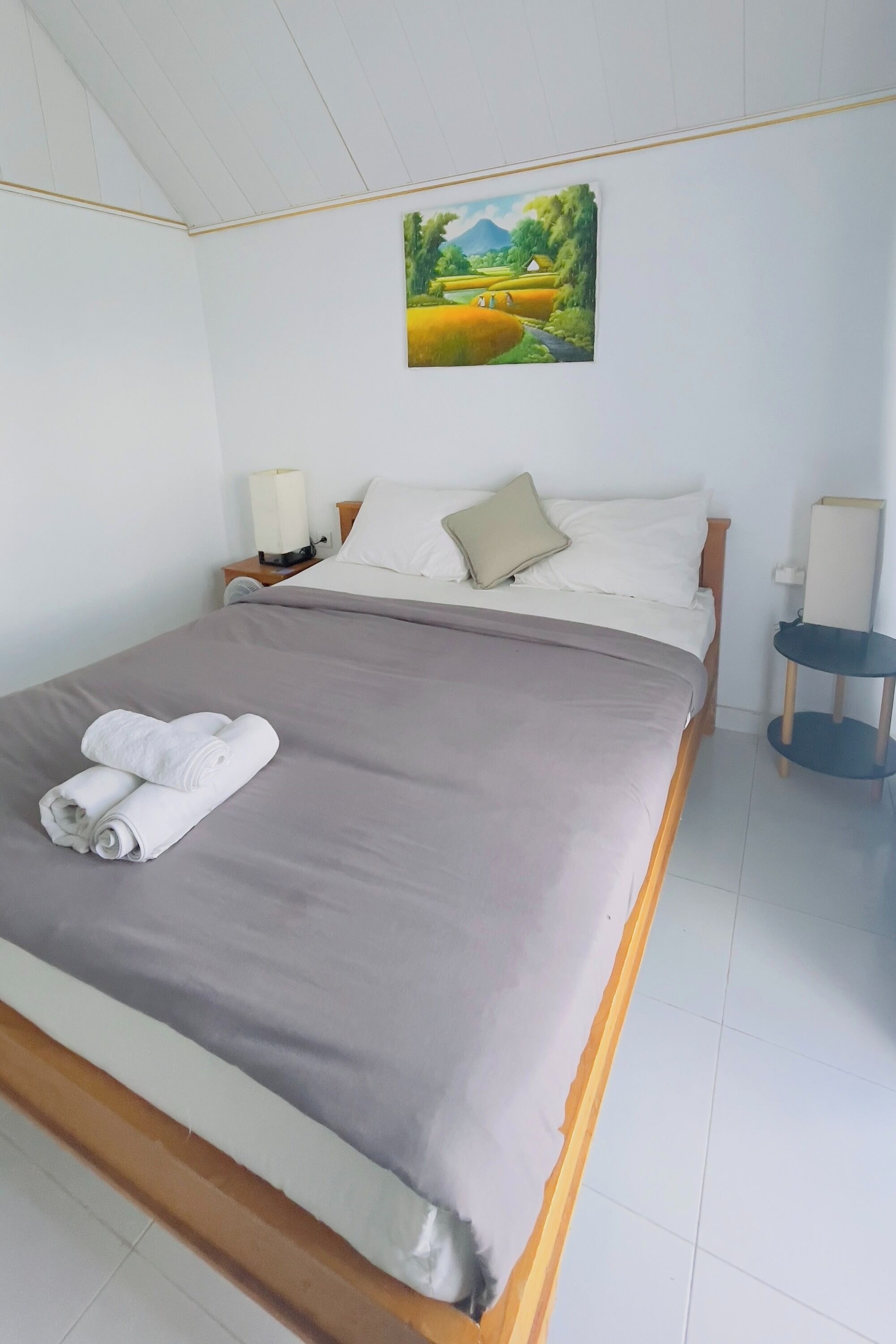 Una cama bien hecha con una colcha gris y dos toallas enrolladas, una mesita de noche con una lámpara y un cuadro de un paisaje en la pared.
