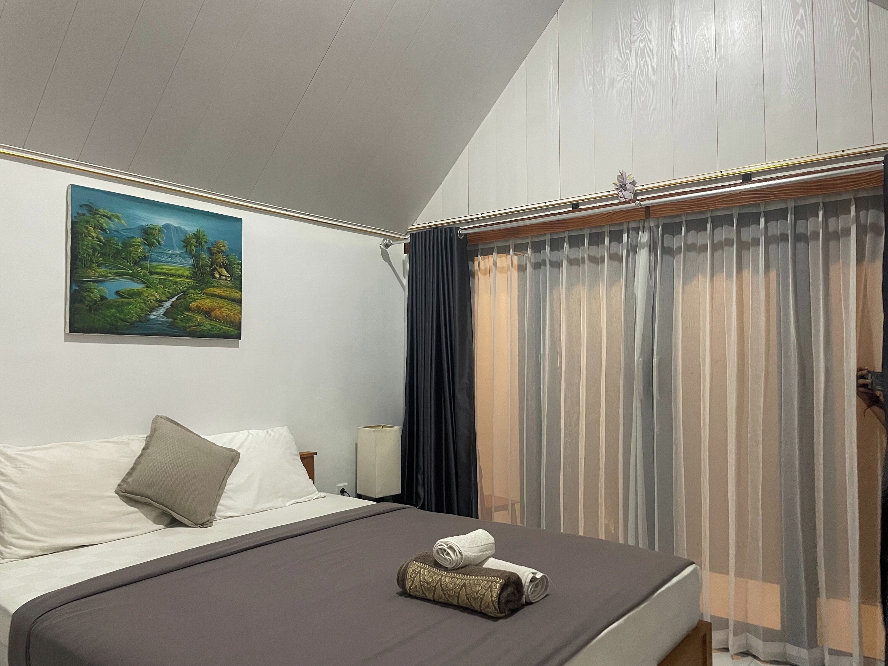 A hotel room with a bed, a painting of a landscape, and a window with sheer curtains.