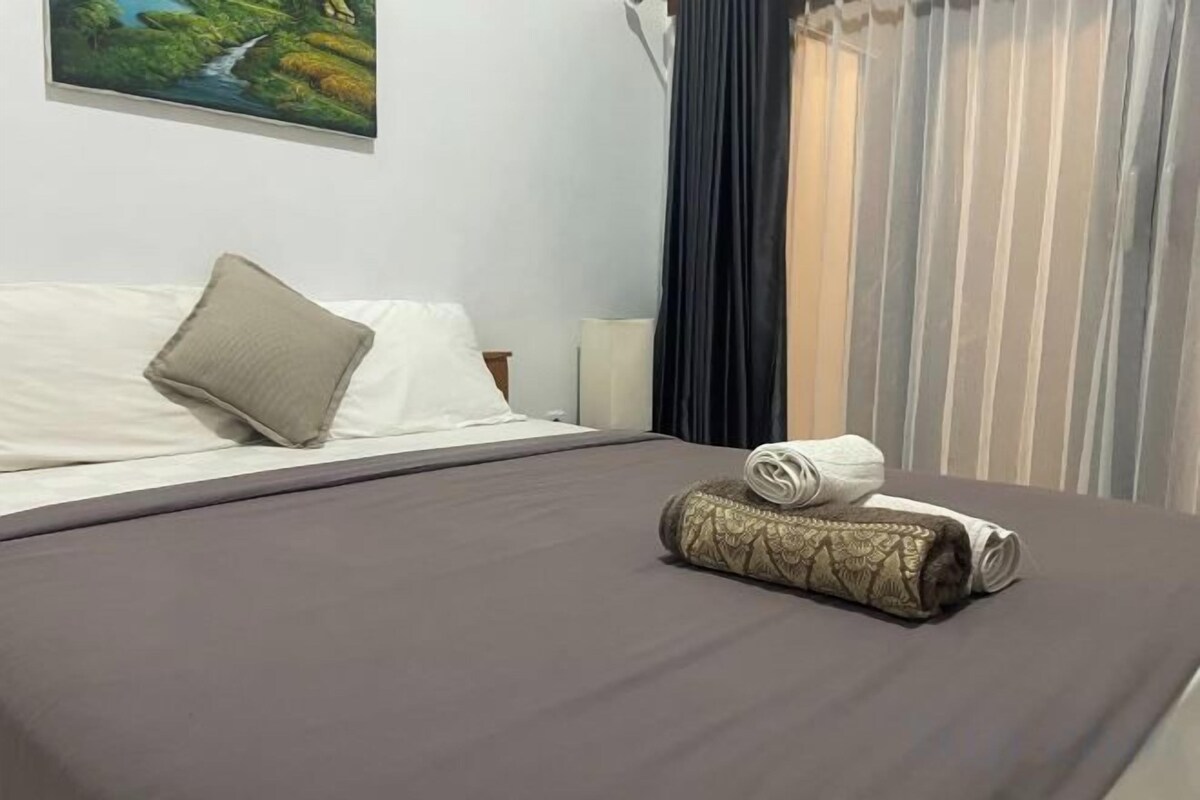 Een net opgemaakt bed met een kussen en opgerolde handdoeken, een nachtkastje met een lamp, en een schilderij aan de muur.