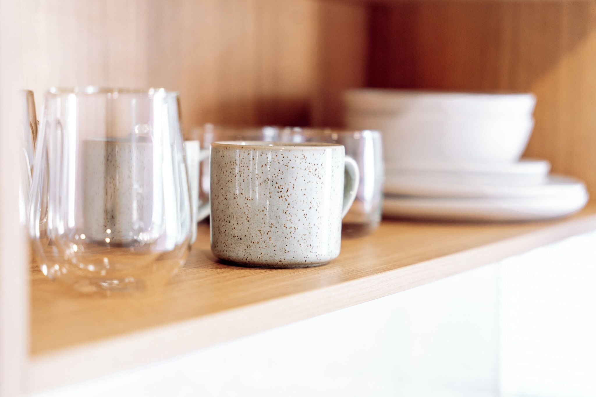 Una encimera de cocina con vasos de vidrio transparente, una taza de cerámica con motas y platos blancos apilados.