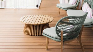 A wooden deck with a round table and two wicker chairs, a white railing, and a view of a pool and indoor area.