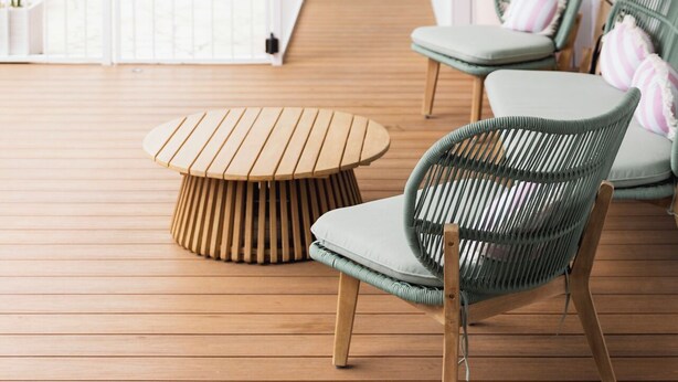 A wooden deck with a round table and two wicker chairs, a white railing, and a view of a pool and indoor area.