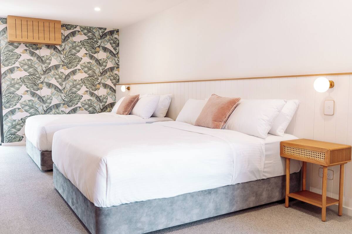 A bedroom with two beds, a wooden bedside table, and a wall with a leaf pattern.