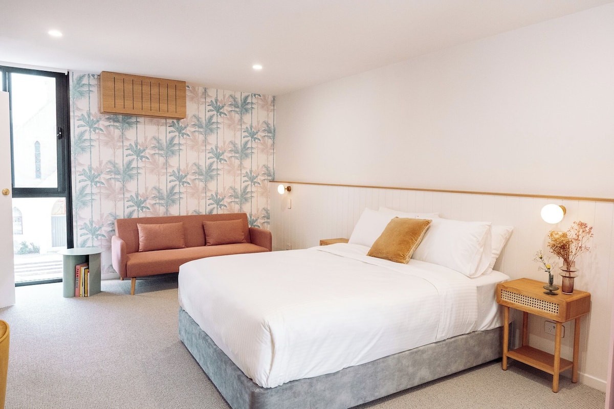 A bedroom with a bed, a sofa, a bedside table with a lamp, and a window.