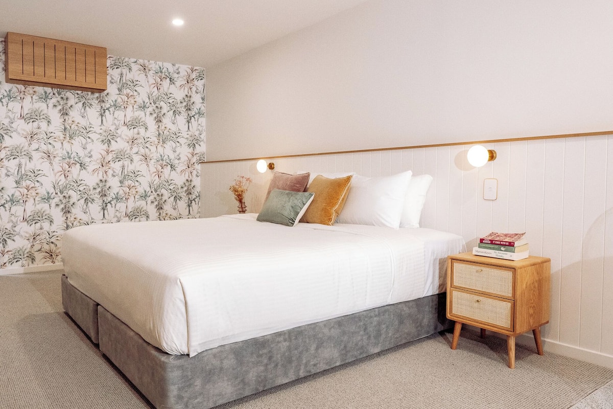 A neatly made bed with a grey base and white linens, a bedside table with a woven basket, and a wall with a tropical wallpaper pattern.