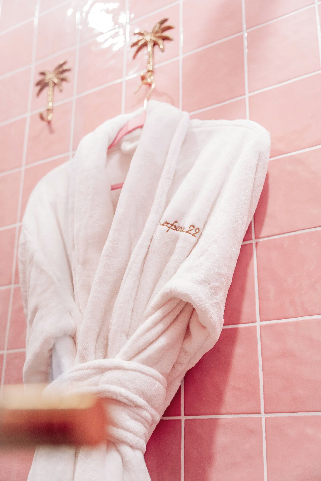 Um roupão branco com um pequeno texto bordado pendurado em uma parede revestida de azulejos rosa.