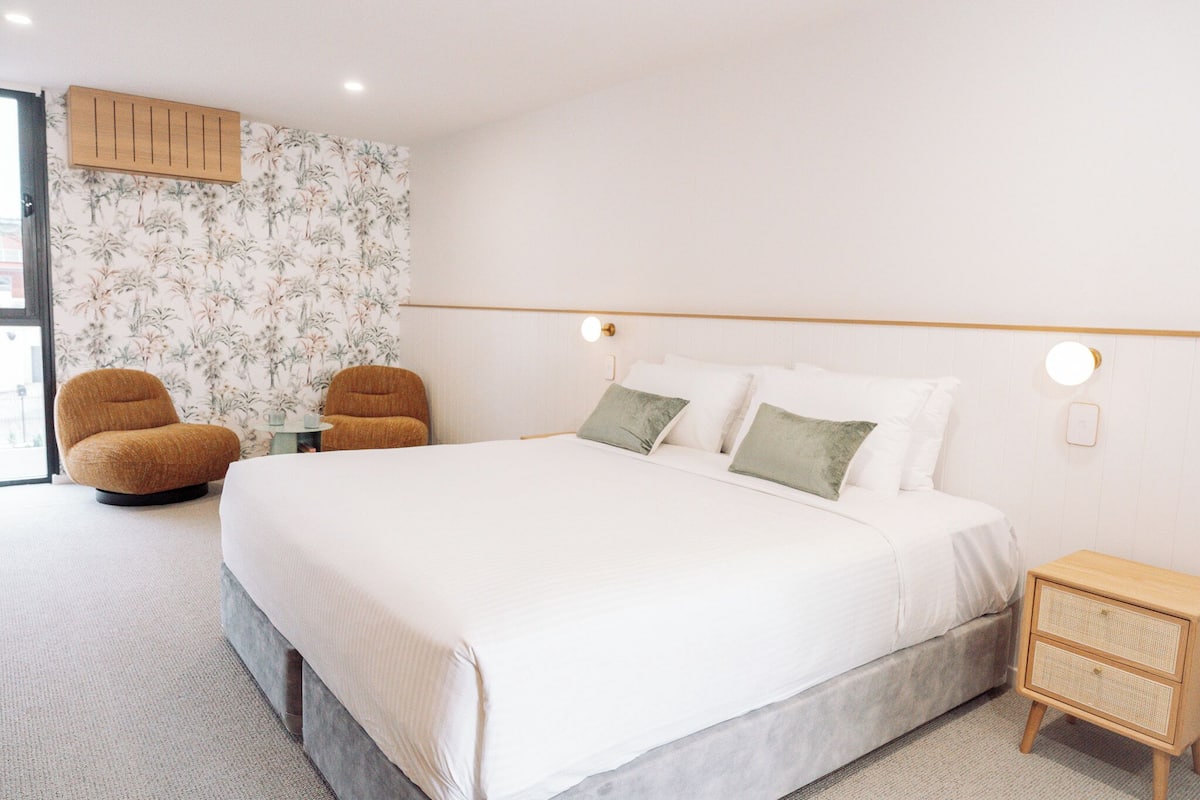 A bedroom with a large bed, two armchairs, a bedside table, and a wall-mounted light fixture.
